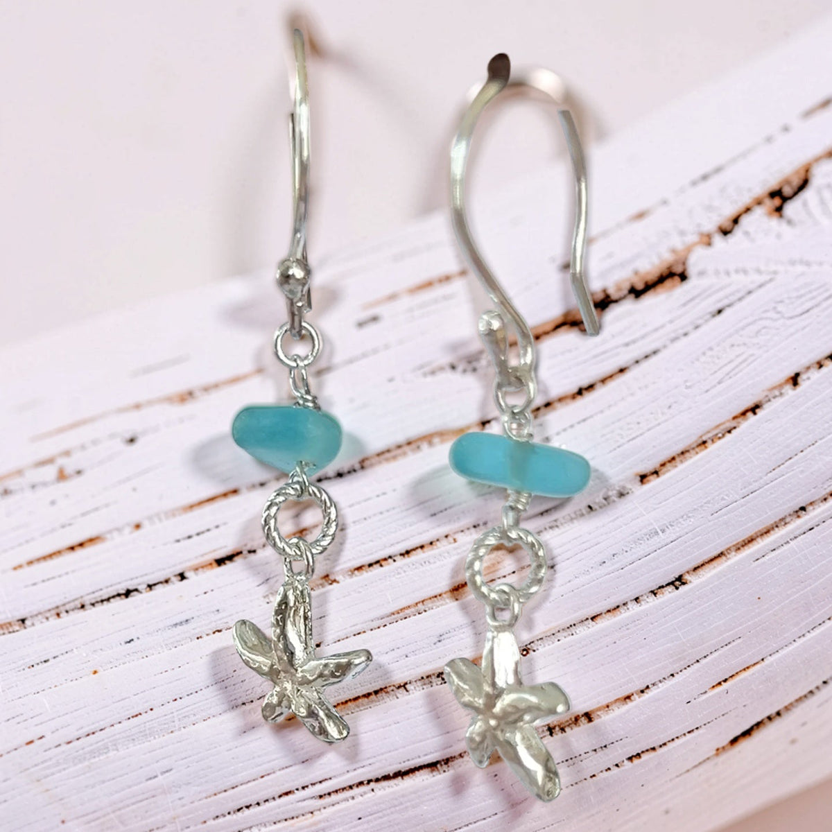 A pair of silver drop earrings with a smooth light blue sea glass bead and a small silver starfish charm, displayed on a white rustic wooden surface.