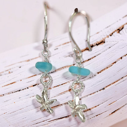 A pair of silver drop earrings with a smooth light blue sea glass bead and a small silver starfish charm, displayed on a white rustic wooden surface.