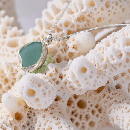 A silver-toned necklace with a smooth, rounded sea glass pendant in a soft mint green color, displayed on a textured white coral branch.
