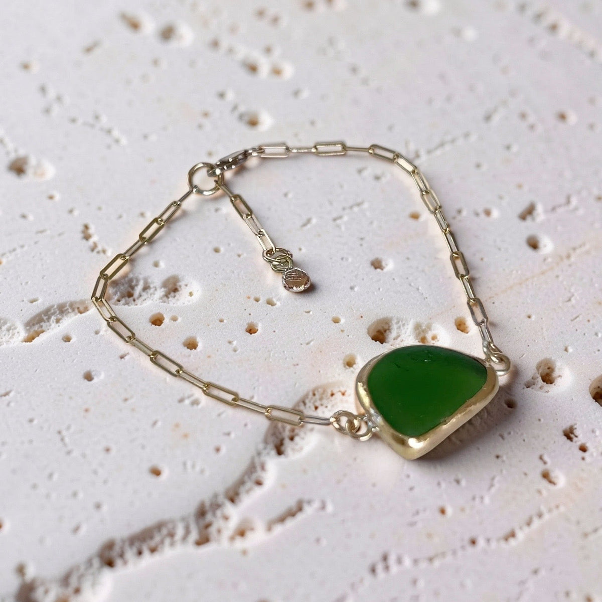 A delicate gold chain bracelet featuring a polished, teardrop-shaped green stone pendant, displayed on a textured, light beige stone surface.