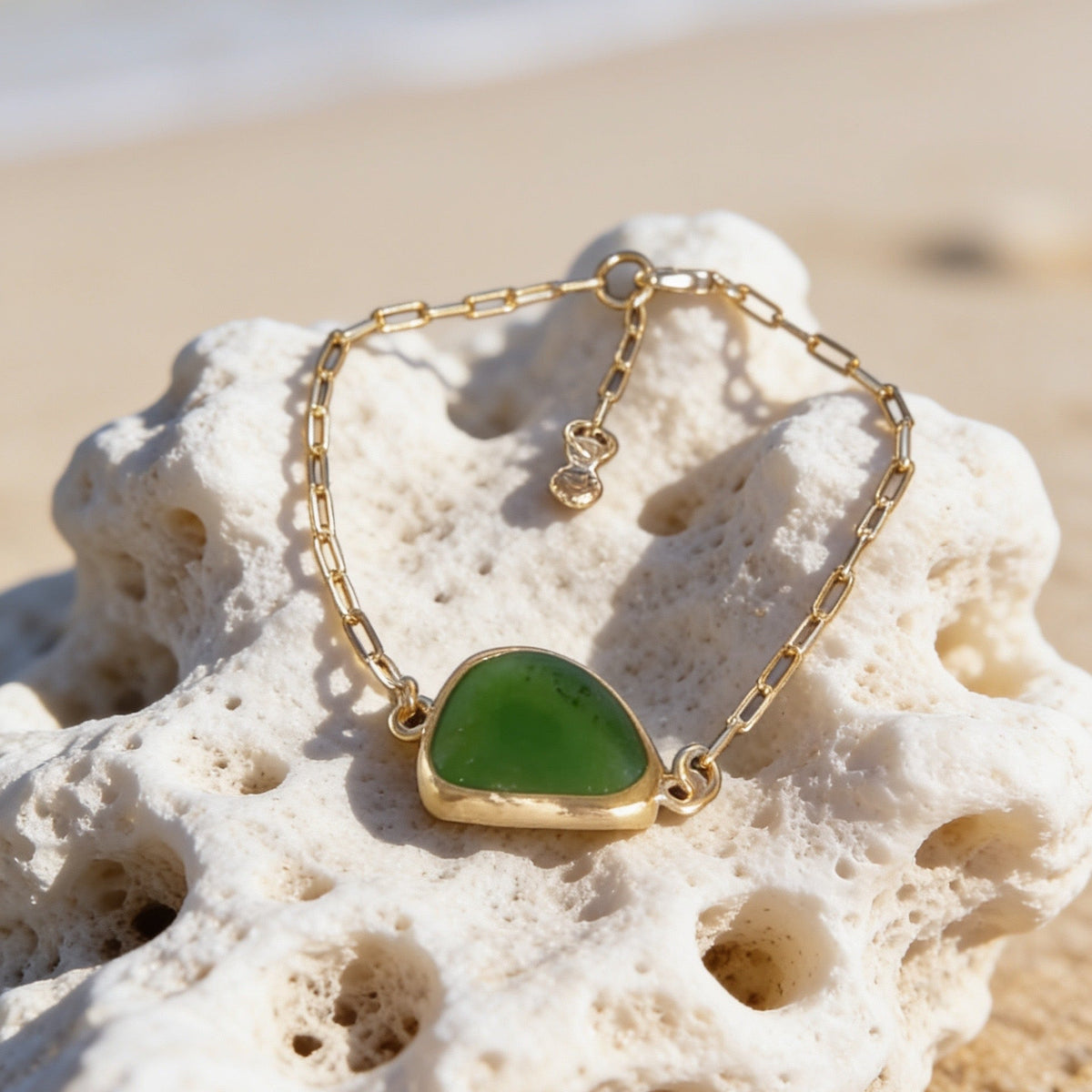 A gold chain bracelet with a polished green sea glass pendant, displayed on a white coral fragment over a sandy beach with the ocean blurred in the background.