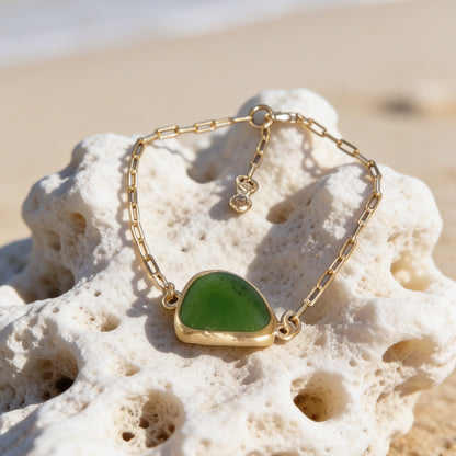 A gold chain bracelet with a polished green sea glass pendant, displayed on a white coral fragment over a sandy beach with the ocean blurred in the background.