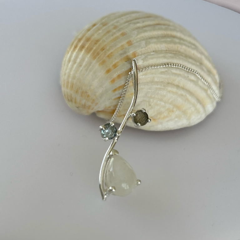 A silver-toned dangle earring with a teardrop-shaped clear stone and a smaller round stone, displayed draped over a beige clam shell on a white plate.