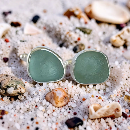 Sea glass ring with two rounded light green stone settings in a silver band, resting on white sand with small shells and pebbles.
