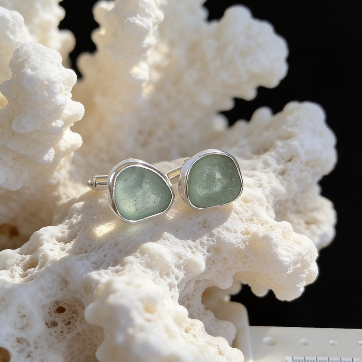 A pair of silver stud earrings with irregular light green stone inlays, displayed on a white coral-like prop against a black background.